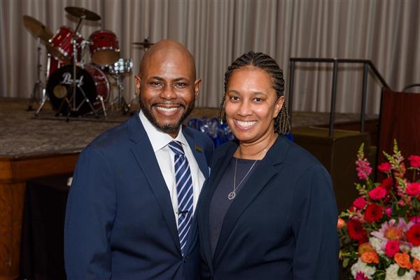 Two people wearing business professional clothing smile at the camera. Behind them, a bouquet of flowers and a stage can be seen. On the stage is a drum set. 